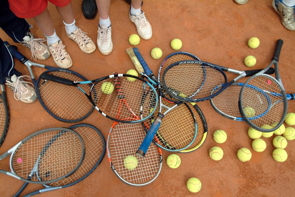 Ansammlung von Tennisschlägern und Bällen auf einem Sandplatz, umgeben von Füßen in Sportschuhen.