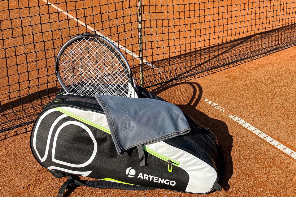 Tennisplatz mit Netz, einer ARTENGO-Tasche und einem Schläger im Vordergrund auf dem rotem Sand.